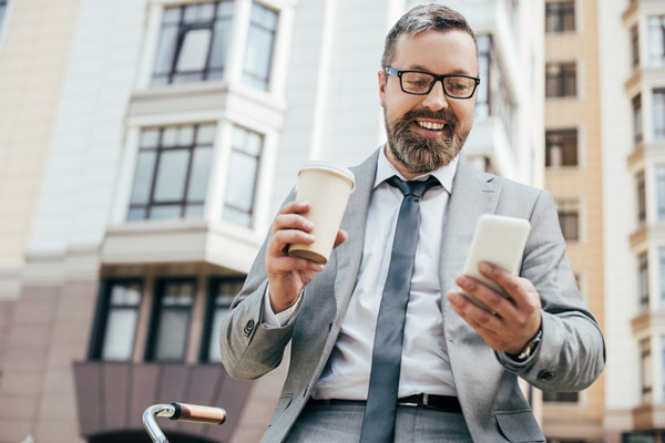 A middle-aged businessman with a beard is smiling while holding a paper coffee cup in one hand and using a smartphone in the other. He is standing outdoors, dressed in a grey suit and tie, with an urban setting and buildings in the background.